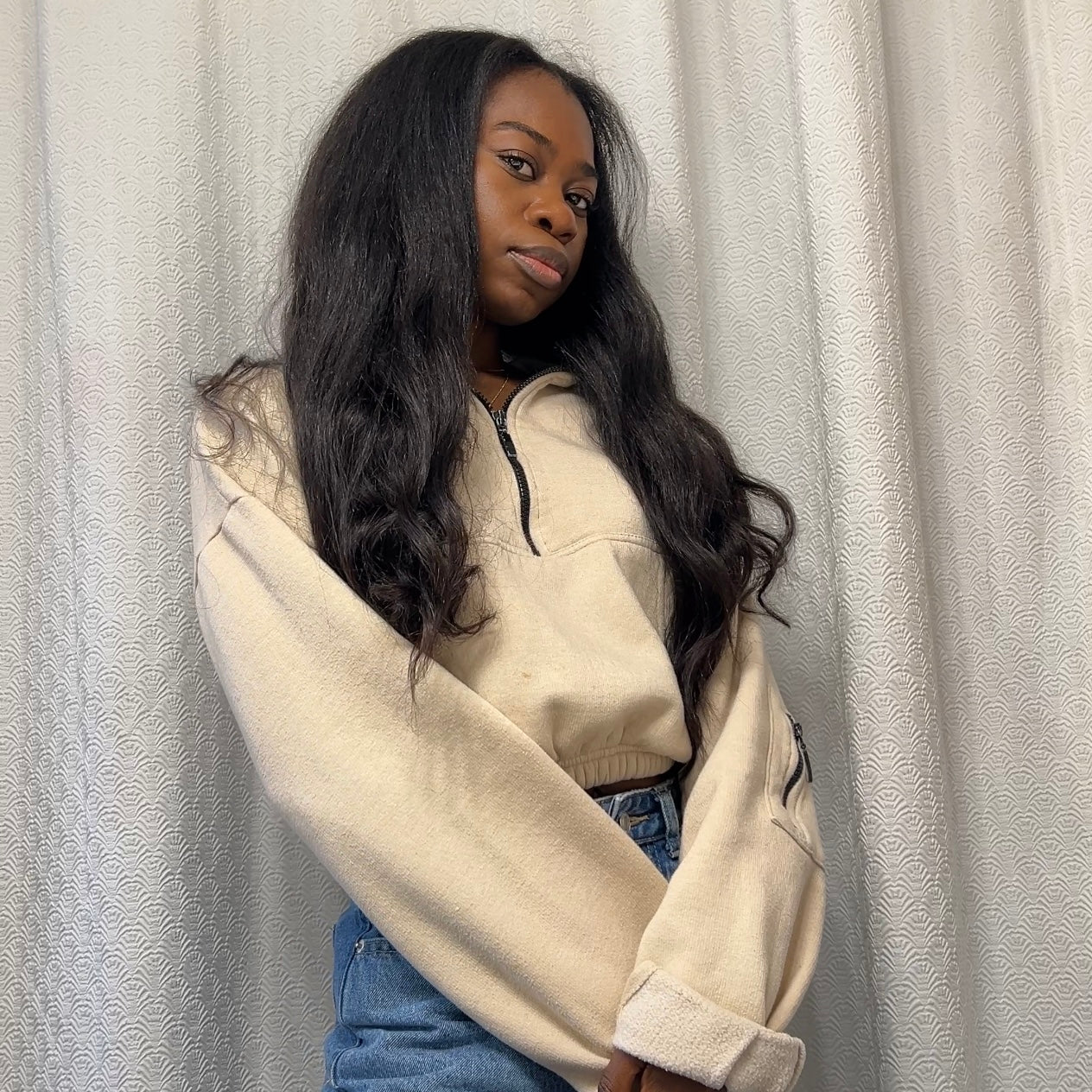 Person wearing a beige hoodie and blue jeans against a plain background