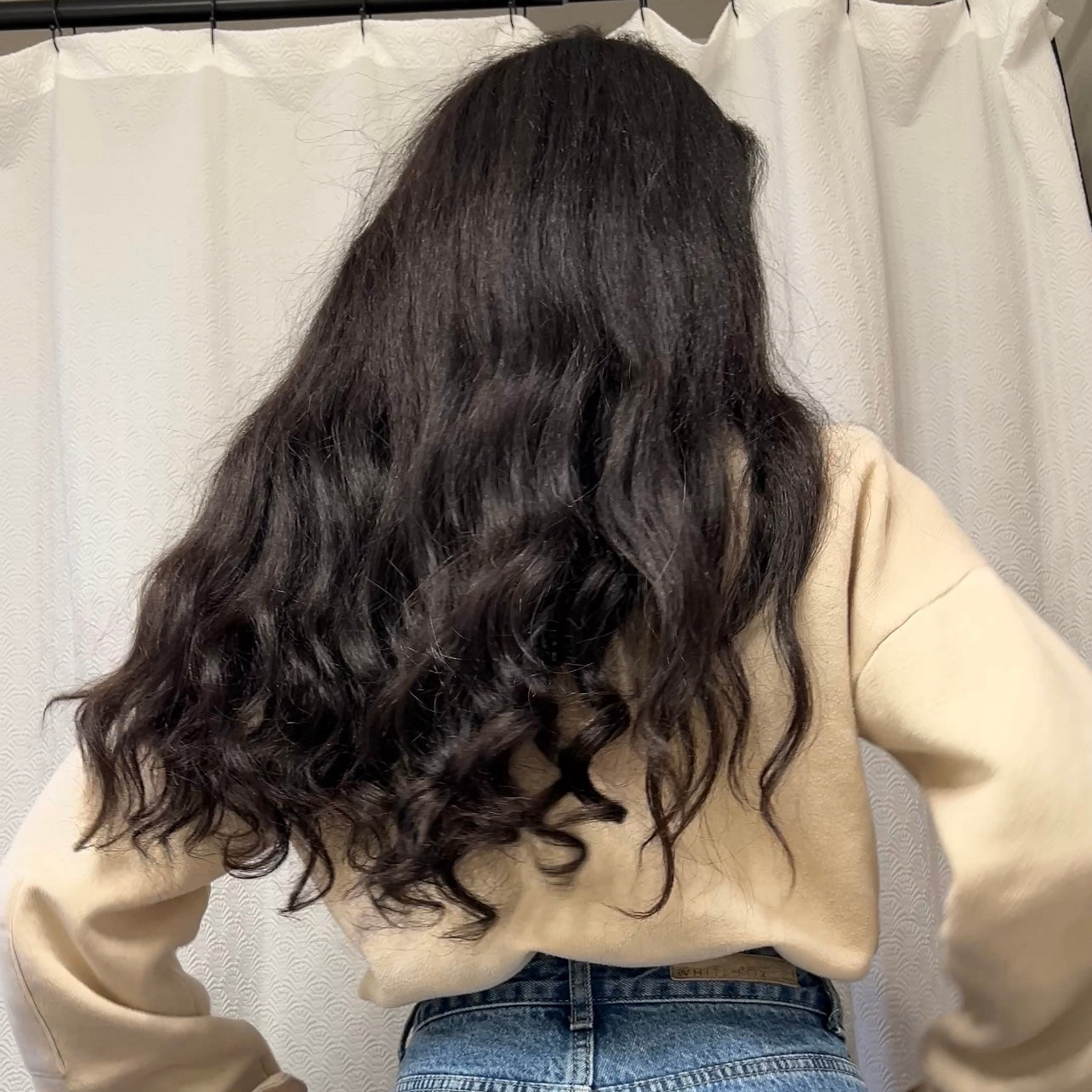 Person with long, wavy black kinky straight hair wearing a beige sweater and blue jeans against a neutral background