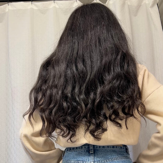 Person with long, wavy black kinky straight hair that has curls at the ends wearing a beige sweater and blue jeans against a plain background